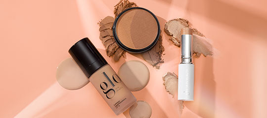 Flat lay of Glo cosmetics: liquid foundation bottle labeled 'glo', pressed powder compact and white lipstick tube on peach background.