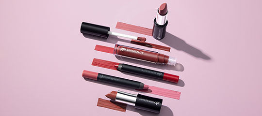 Assorted mauve and nude lipsticks and glosses arranged diagonally on a pale pink background with color swatches.