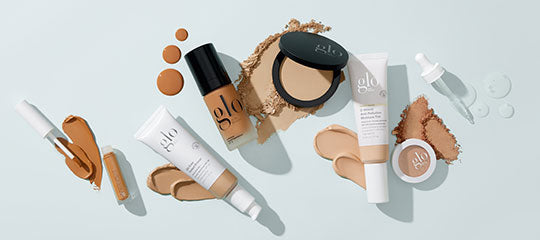 Assorted glo cosmetic products with foundation swatches—tubes, compact, bottle, dropper, and scattered powder on pale blue background.