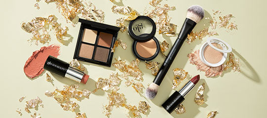 Makeup flat-lay with eyeshadow quad, glo powder compact, brush, bareMinerals lipstick and cream blush with gold leaf flakes.