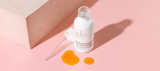 Bottle of glo serum with glass dropper and two orange serum drops on pink background