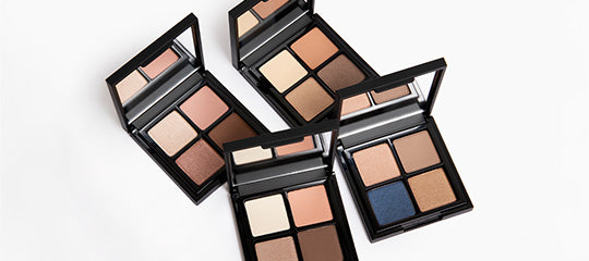 Four open eyeshadow palettes showing neutral and blue shimmer shades in square pans with built-in mirrors.