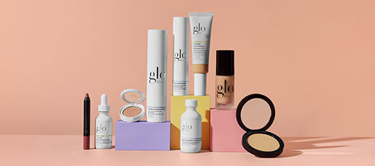 Assorted glo skincare and makeup bottles, compact powder, and lipstick displayed on colored blocks against a peach background