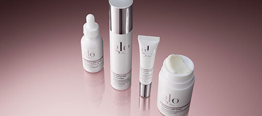 Four white glo skincare containers: dropper bottle, pump bottle, tube, and open jar on a reflective pink surface
