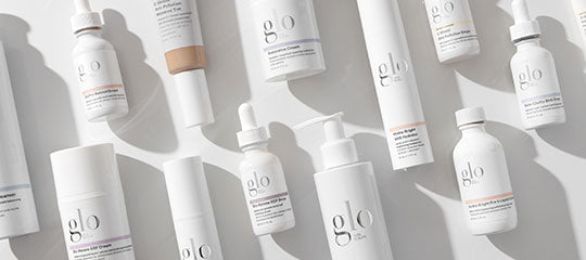 Assorted white glo skincare bottles, dropper and pump dispensers, jars and tubes arranged on a pale background.