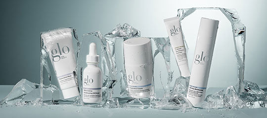 Glo skincare lineup on cracked ice: five white glo-branded tubes, dropper bottle and jar arranged on ice shards