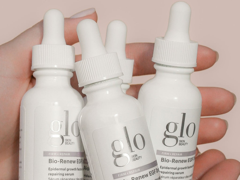 Hand holding three white glo SKIN BEAUTY bottles labeled Bio-Renew EGF FIRM + REPAIR Epidermal growth factor repairing serum
