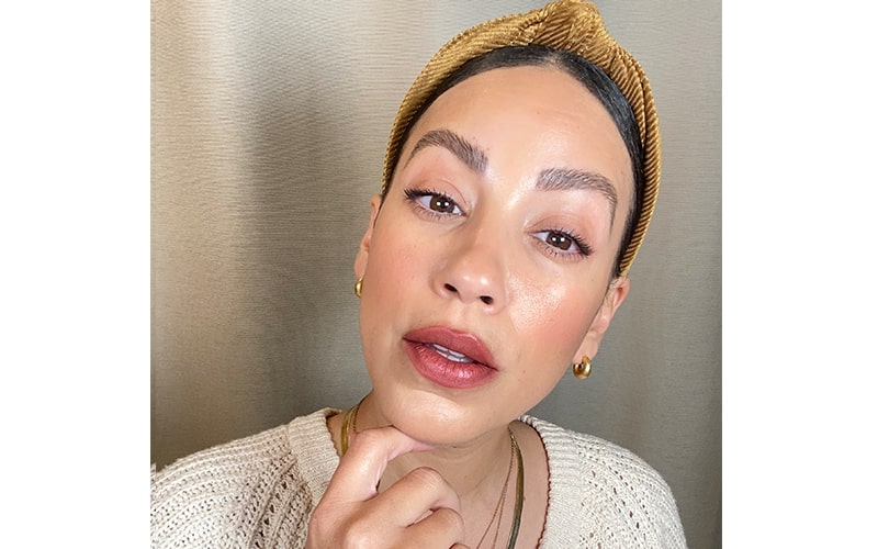 Close-up of a woman with wine-stained lipstick, gold hoop earrings and headband, cream knit sweater, hand resting on chin.