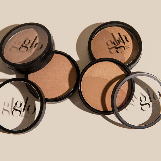 Multiple open bronze powder compacts on beige background with glo SKIN BEAUTY printed on lids