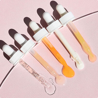 Five cosmetic droppers with white caps holding clear, pink, peach, orange, and cream serums on a pink background