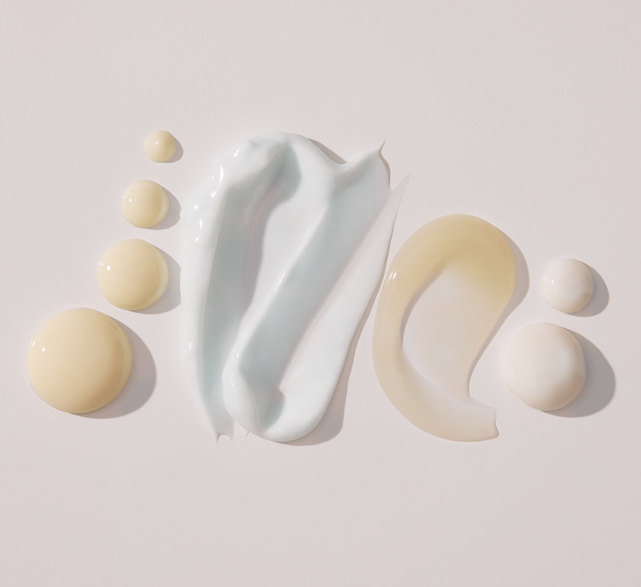 Skincare swatches: pale blue cream smear center, translucent beige gel smear right, and several round pale-yellow droplets.