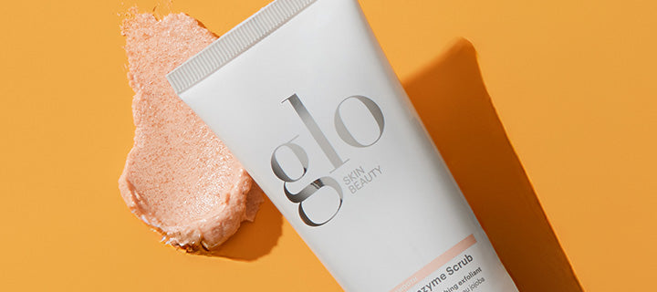 Tilted white tube labeled glo SKIN BEAUTY Enzyme Scrub with peach scrub smear on orange background