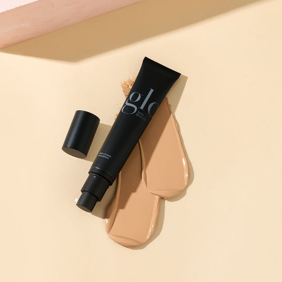 Black tube with glo SKIN BEAUTY logo and text Satin Cream Foundation, on two beige foundation swatches