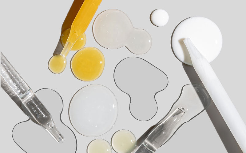 Glass and plastic droppers and scattered serum droplets (clear, white, yellow) on a gray background