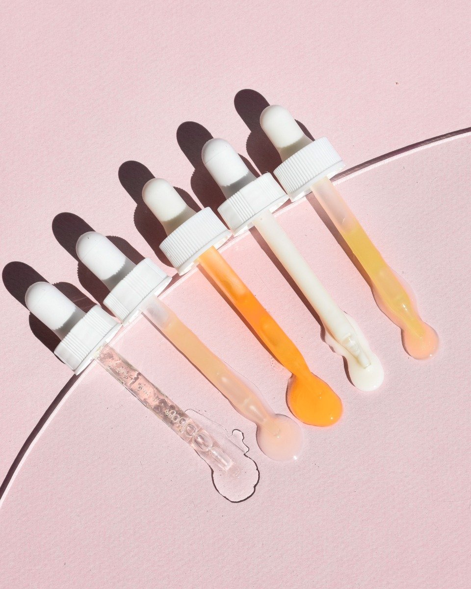 Five white-topped droppers leaking clear, peach, orange, and ivory serums onto a curved pale-pink surface