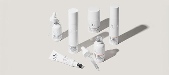 Assorted white glo skincare bottles and dropper lying on a pale surface with long shadows