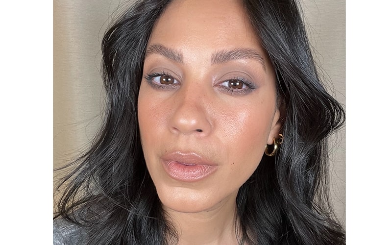 Close-up of a woman with cool-toned smoky eye makeup, glossy nude lips, wavy dark hair and small gold hoop earring.