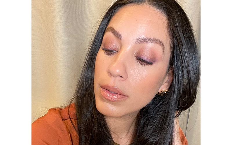 Woman with closed eyes showing copper smokey-eye makeup, glossy lips and dark hair tucked behind ear