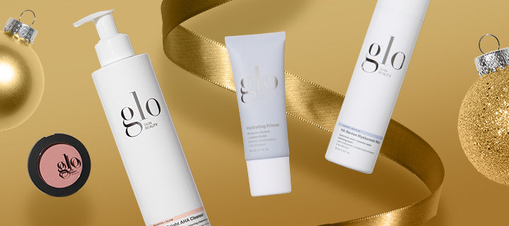 Three glo skin beauty products — pump bottle, Hydrating Primer tube, and cylinder bottle on gold background with ribbon and ornaments.