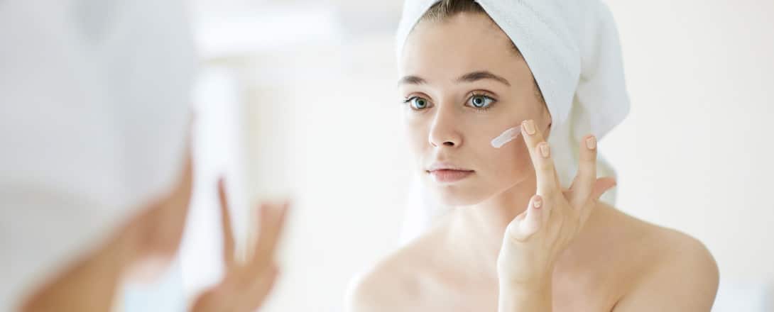 skincare-hero Photo of young woman applying skincare products to face while looking in the mirror.